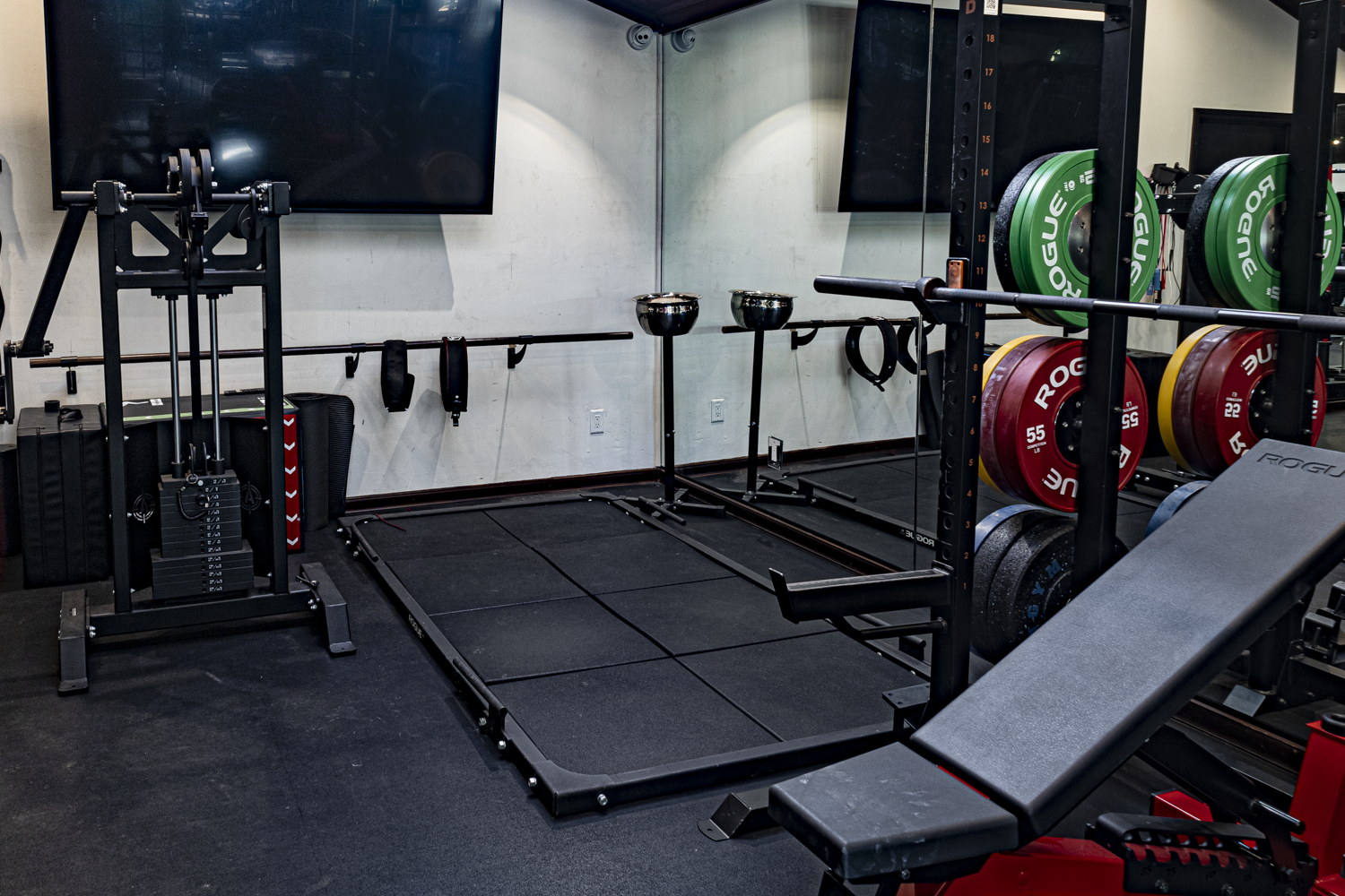 Weightlifting platform with Rogue rack, color-coded bumper plates, cable machine, and wall-mounted screens