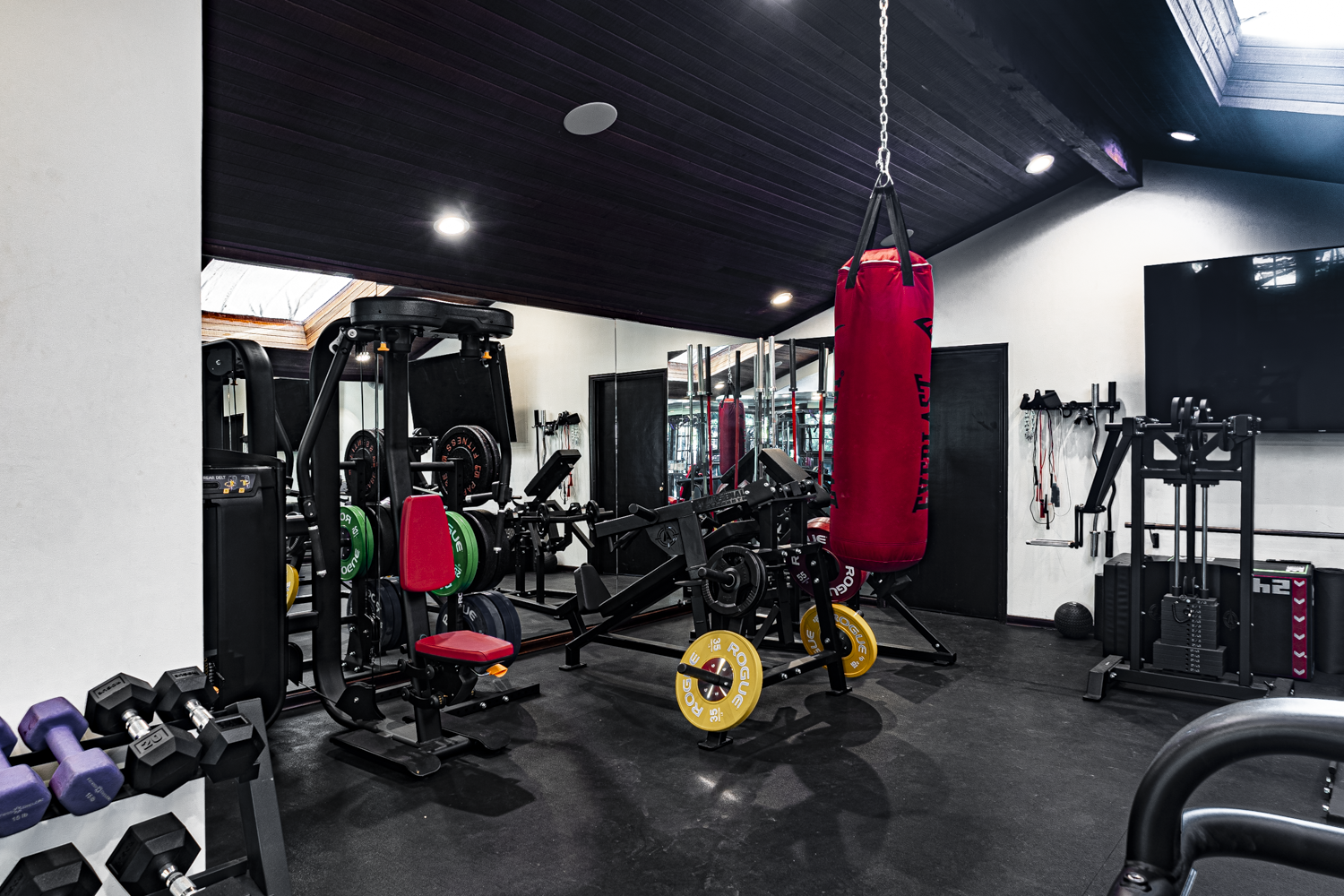 Plate-loaded machines, leg press, and heavy bag under a dark wood ceiling with a skylight