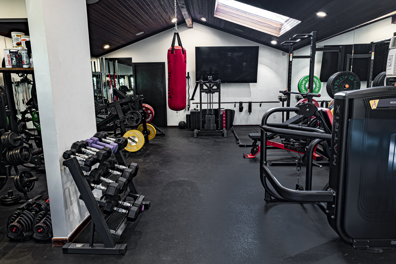 Wide view of a home gym with dumbbell rack, heavy bag, bench, and Matrix machine under a vaulted wood ceiling