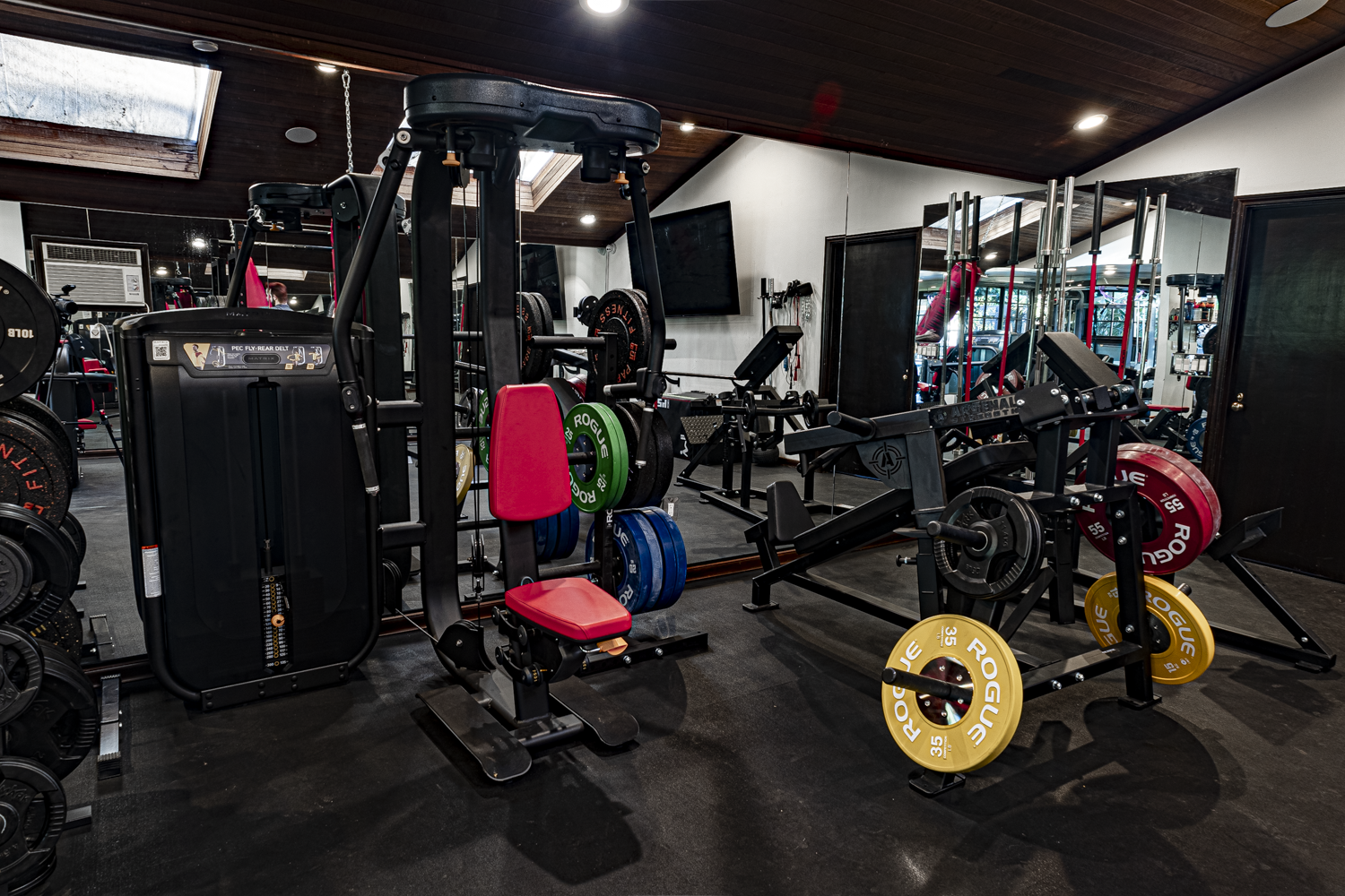 Pec fly machine, Arsenal Strength press, and color-coded Rogue plates under a wood-paneled ceiling with skylights